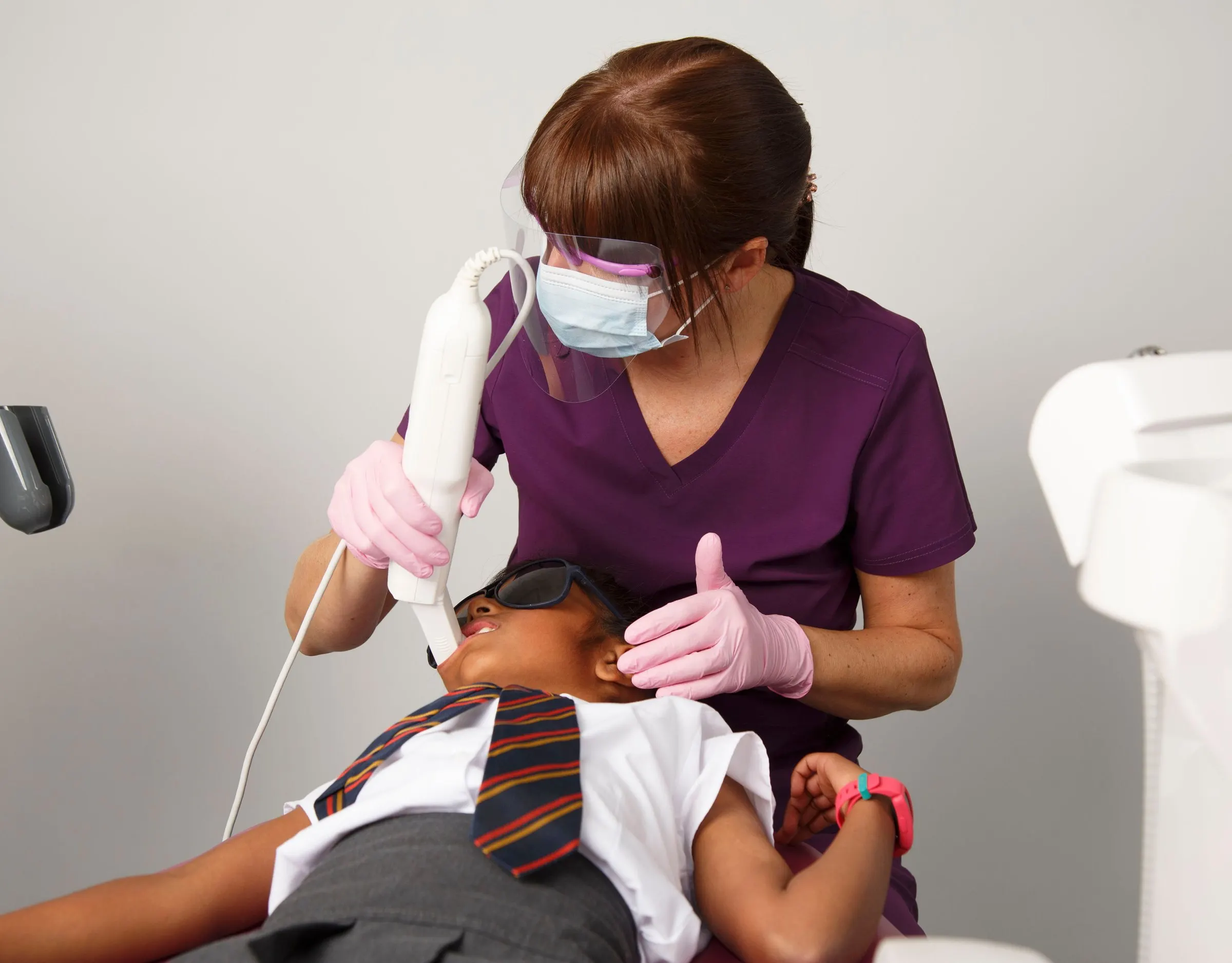 A clinician in purple scrubs, mask and visor treating a young patient in the dental chair